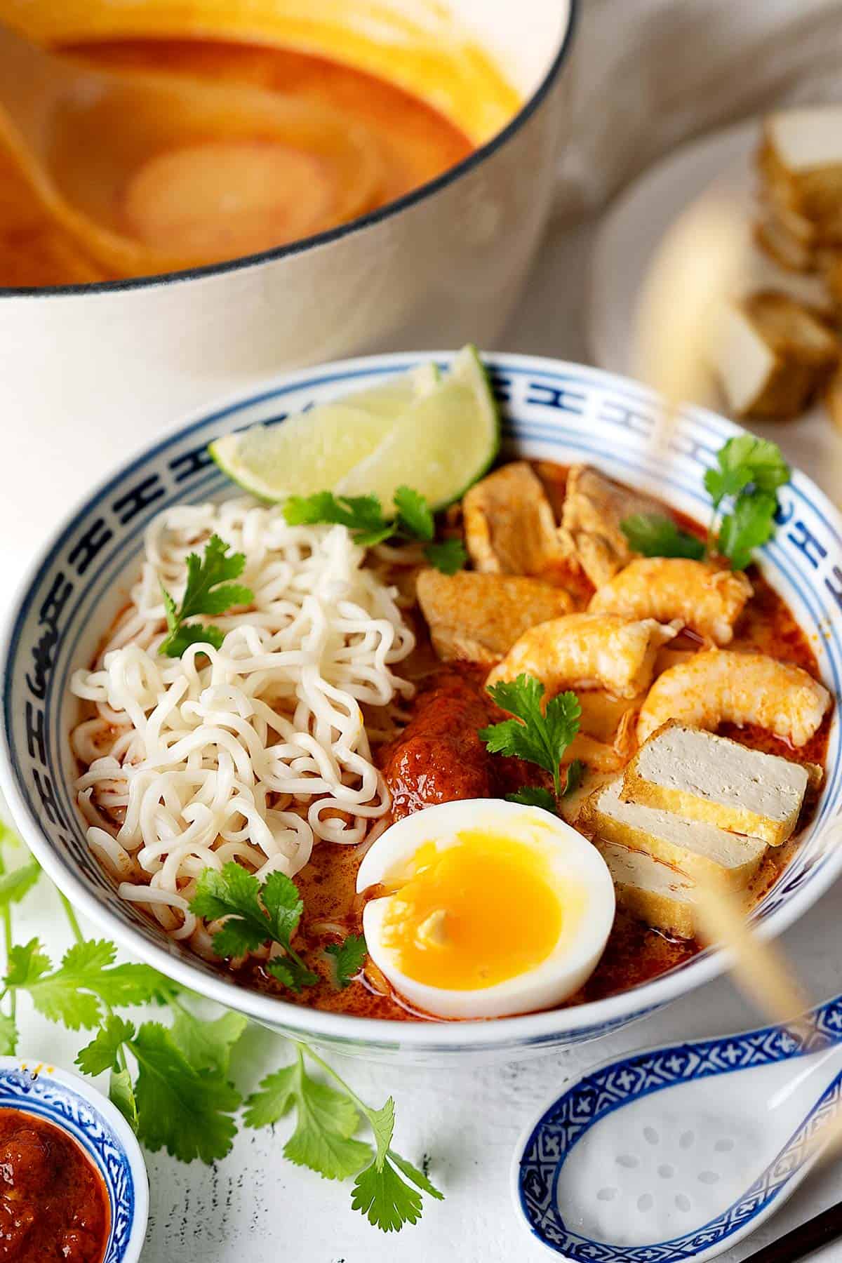 A bowl of quick red curry laksa with a pot at the back and condiments on the side