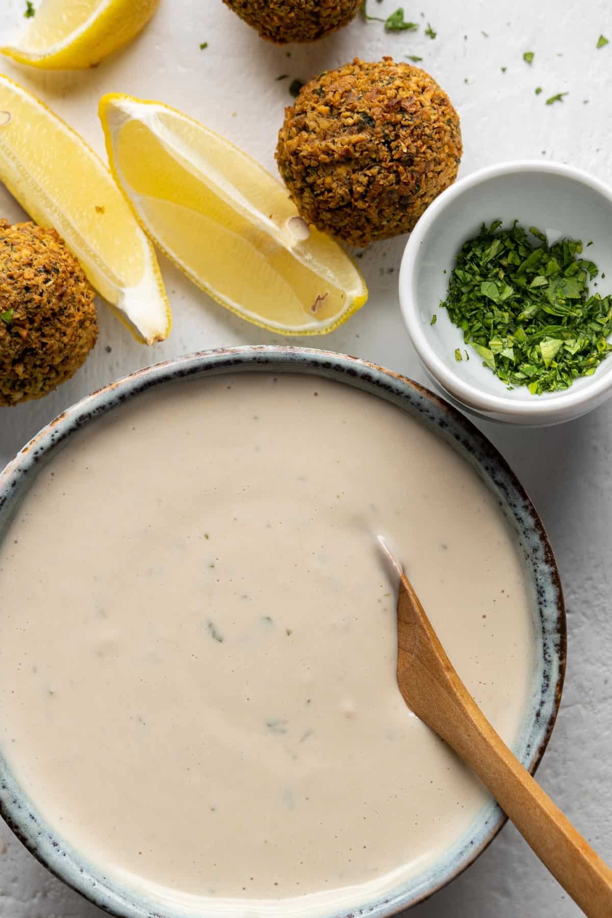 Tahini sauce in a bowl with some falafel and lemon wedges.
