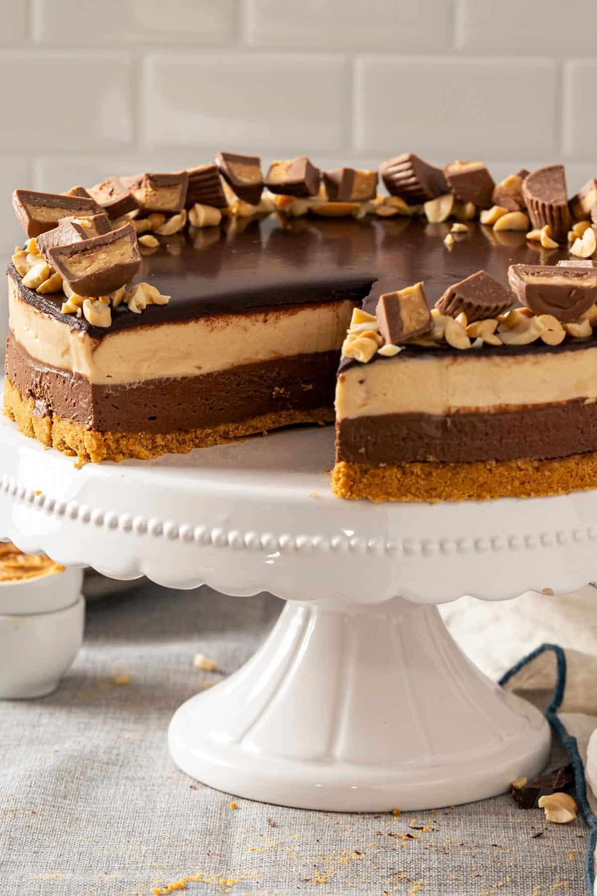 Whole no bake peanut butter chocolate cheesecake on a white stand topped with ganache and peanut butter cups with a slice missing.