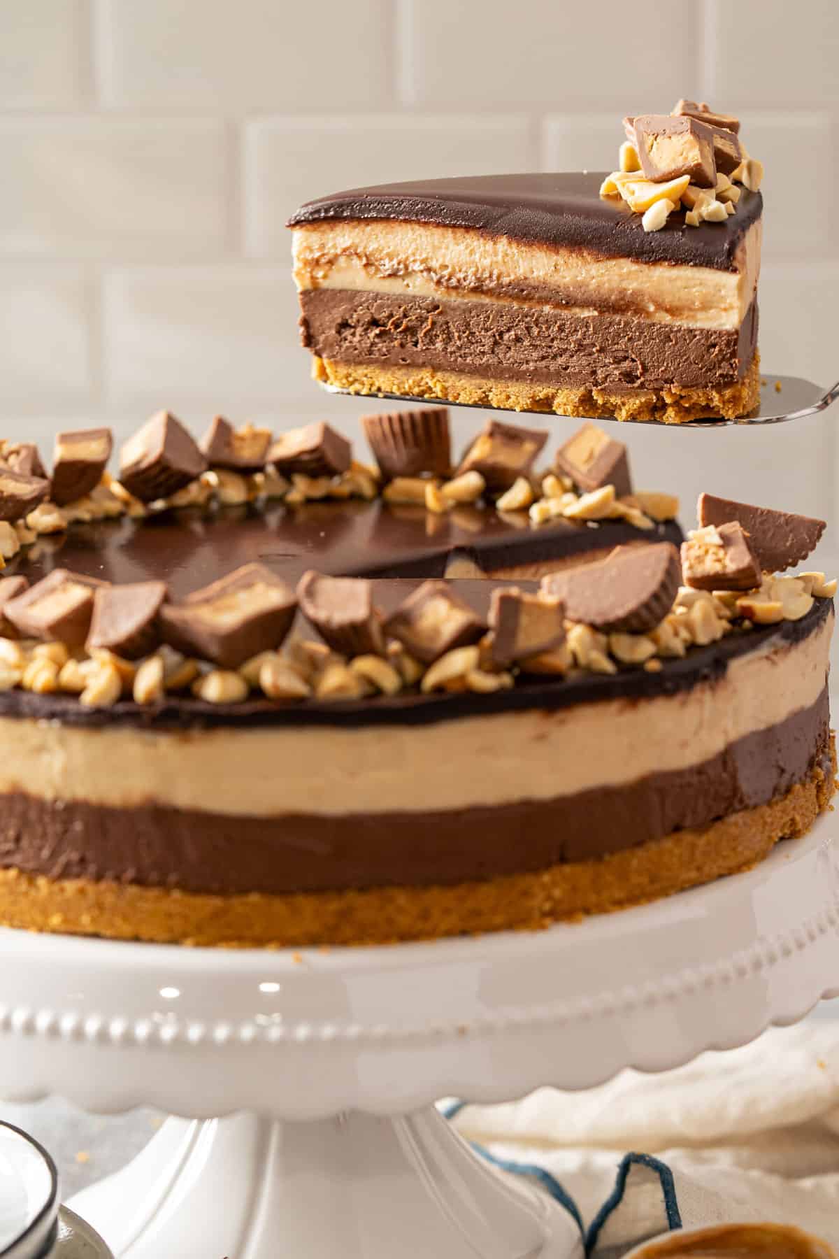 Lifting a slice of no bake peanut butter chocolate cheesecake on a white stand topped with ganache and peanut butter cups.