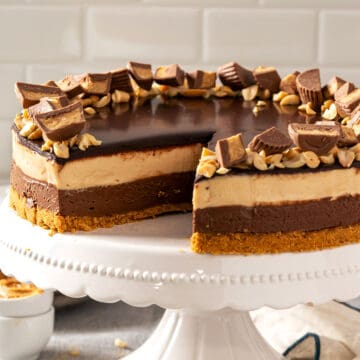 Whole no bake peanut butter chocolate cheesecake on a white stand topped with ganache and peanut butter cups.