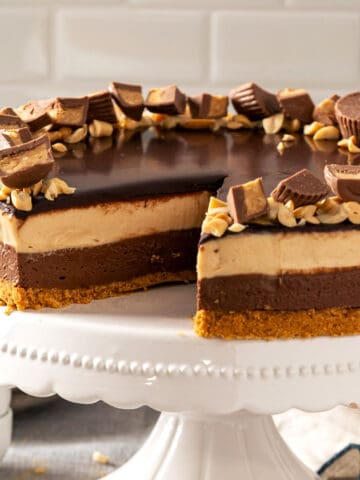Whole no bake peanut butter chocolate cheesecake on a white stand topped with ganache and peanut butter cups.
